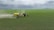 A green tractor is towing a large yellow agricultural sprayer across a vast field of crops under an overcast sky. Dust is visible behind the equipment, indicating movement and activity in tending to the field.
