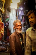 A market scene with two men; one with a gray beard wearing a dark jacket and shirt, standing under bright lights. Hanging meat and various market items are visible in the background, contributing to a busy atmosphere.