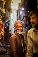 A market scene with two men; one with a gray beard wearing a dark jacket and shirt, standing under bright lights. Hanging meat and various market items are visible in the background, contributing to a busy atmosphere.