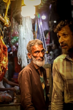 A market scene with two men; one with a gray beard wearing a dark jacket and shirt, standing under bright lights. Hanging meat and various market items are visible in the background, contributing to a busy atmosphere.