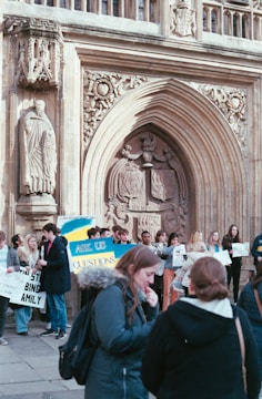 A group of people gathered outside an ornate, historical building, holding signs and engaging in conversation. The architecture features detailed carvings and an arched doorway with a statue. Some signs are visible, including one that says 'ASK US QUESTIONS'. The atmosphere suggests a casual, informative event or protest.