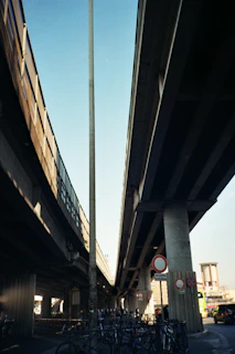 Two large concrete highways or overpasses stretch above, with multiple street signs visible below. A bicycle parking area is located underneath, with several bikes attached to metal racks. A tall streetlamp and urban infrastructure, including buildings in the distance, provide a cityscape background. Soft shadows are cast by the structures above, indicating sunlight.