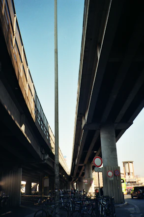 Two large concrete highways or overpasses stretch above, with multiple street signs visible below. A bicycle parking area is located underneath, with several bikes attached to metal racks. A tall streetlamp and urban infrastructure, including buildings in the distance, provide a cityscape background. Soft shadows are cast by the structures above, indicating sunlight.