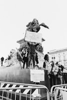 A group of people gathered around a monument are holding protest signs. One sign prominently reads, 'Russian warship go f*ck yourself,' while another held by a child reads, 'Peace for Ukraine.' The atmosphere appears tense but hopeful. The scene is black and white, adding a dramatic and serious tone to the protest.
