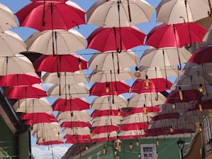 Rows of red and white umbrellas hanging upside down form a canopy across an outdoor alley, creating a playful and vibrant visual display. Above, the blue sky is visible in between the gaps. Light bulbs are interspersed between the umbrellas, suggesting a decorative or artistic installation.