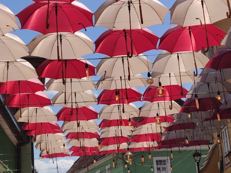 Rows of red and white umbrellas hanging upside down form a canopy across an outdoor alley, creating a playful and vibrant visual display. Above, the blue sky is visible in between the gaps. Light bulbs are interspersed between the umbrellas, suggesting a decorative or artistic installation.