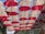 Rows of red and white umbrellas hanging upside down form a canopy across an outdoor alley, creating a playful and vibrant visual display. Above, the blue sky is visible in between the gaps. Light bulbs are interspersed between the umbrellas, suggesting a decorative or artistic installation.