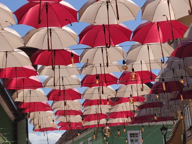 Rows of red and white umbrellas hanging upside down form a canopy across an outdoor alley, creating a playful and vibrant visual display. Above, the blue sky is visible in between the gaps. Light bulbs are interspersed between the umbrellas, suggesting a decorative or artistic installation.