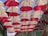 Rows of red and white umbrellas hanging upside down form a canopy across an outdoor alley, creating a playful and vibrant visual display. Above, the blue sky is visible in between the gaps. Light bulbs are interspersed between the umbrellas, suggesting a decorative or artistic installation.