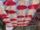 Rows of red and white umbrellas hanging upside down form a canopy across an outdoor alley, creating a playful and vibrant visual display. Above, the blue sky is visible in between the gaps. Light bulbs are interspersed between the umbrellas, suggesting a decorative or artistic installation.