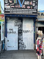 A shop's metal shutter features graffiti art with a realistic depiction of a face peering through fingers. The phrase 'BE CURIOUS... ALWAYS!' is written in large letters. The storefront is surrounded by various colorful garments displayed on mannequins, and the shop sign above advertises items like hats, clothing, and accessories.