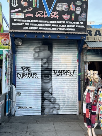 A shop's metal shutter features graffiti art with a realistic depiction of a face peering through fingers. The phrase 'BE CURIOUS... ALWAYS!' is written in large letters. The storefront is surrounded by various colorful garments displayed on mannequins, and the shop sign above advertises items like hats, clothing, and accessories.