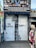A shop's metal shutter features graffiti art with a realistic depiction of a face peering through fingers. The phrase 'BE CURIOUS... ALWAYS!' is written in large letters. The storefront is surrounded by various colorful garments displayed on mannequins, and the shop sign above advertises items like hats, clothing, and accessories.
