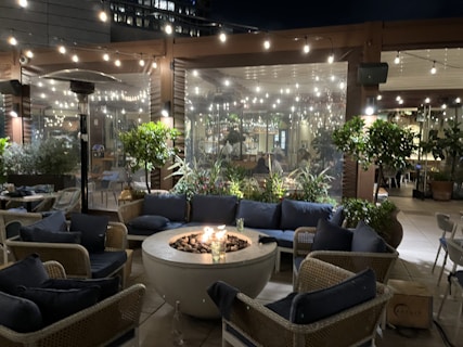 A cozy outdoor seating area is arranged around a round fire pit. The space is adorned with string lights creating a warm and inviting atmosphere. The furniture includes wicker chairs and blue cushions, surrounded by green plants and a backdrop of a glass window, showing people inside the adjacent area.