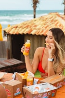 A woman in a red bikini top enjoys snacks and a bottle of orange juice while sitting at a wooden table by the beach. The table has several takeaway food boxes, and the background features a rustic hut with a tiled roof and palm trees near the ocean.