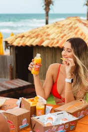 A woman in a red bikini top enjoys snacks and a bottle of orange juice while sitting at a wooden table by the beach. The table has several takeaway food boxes, and the background features a rustic hut with a tiled roof and palm trees near the ocean.