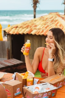 A woman in a red bikini top enjoys snacks and a bottle of orange juice while sitting at a wooden table by the beach. The table has several takeaway food boxes, and the background features a rustic hut with a tiled roof and palm trees near the ocean.