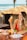 A woman in a red bikini top enjoys snacks and a bottle of orange juice while sitting at a wooden table by the beach. The table has several takeaway food boxes, and the background features a rustic hut with a tiled roof and palm trees near the ocean.