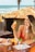 A woman in a red bikini top enjoys snacks and a bottle of orange juice while sitting at a wooden table by the beach. The table has several takeaway food boxes, and the background features a rustic hut with a tiled roof and palm trees near the ocean.
