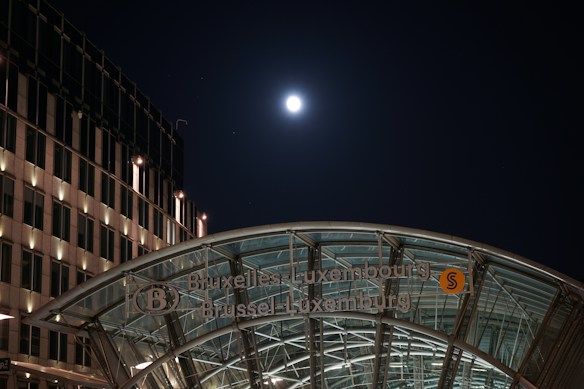 A nighttime urban scene featuring a modern glass and steel structure of a train station entrance with the name 'Bruxelles-Luxembourg' prominently displayed. The moon shines brightly in the dark sky, creating a striking contrast against the illuminated building facade on the left.