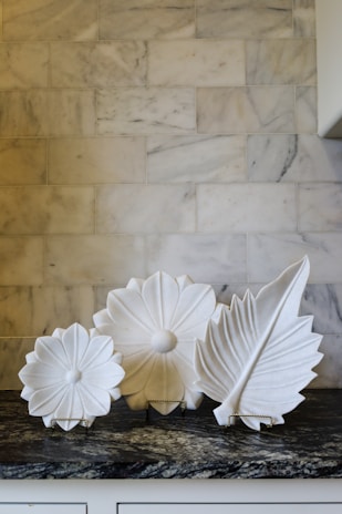 Three decorative white ceramic plates displaying intricate floral and leaf designs are arranged on a patterned marble counter. The background features a light-colored marble tile wall.