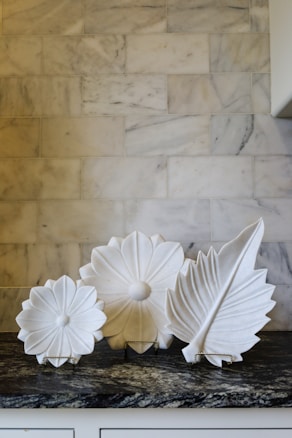 Three decorative white ceramic plates displaying intricate floral and leaf designs are arranged on a patterned marble counter. The background features a light-colored marble tile wall.