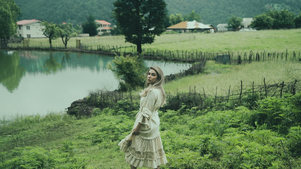 a person in a white dress standing in a field of grass by a pond