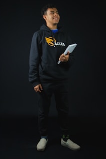 A confident Indian student in a vibrant purple hoodie, smiling while holding textbooks against a bright white background.