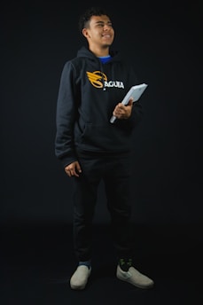 A confident Indian student in a vibrant purple hoodie, smiling while holding textbooks against a bright white background.