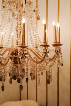 A luxurious chandelier adorned with glistening crystals hanging elegantly, with several candle-shaped light bulbs emitting a warm glow. The background features vertical gold stripes on an off-white wall.