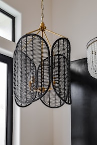 A stylish chandelier made of black woven rattan hangs from the ceiling, featuring a unique design with a central brass fixture. It is surrounded by a modern interior with a mix of soft and dark colors, including nearby elements like large windows and another more decorative light fixture with white beads.