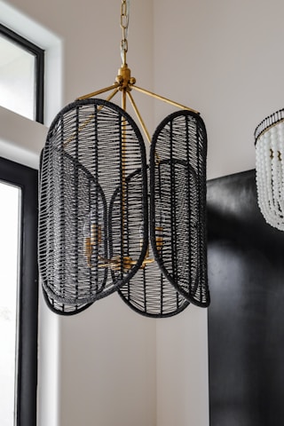 A stylish chandelier made of black woven rattan hangs from the ceiling, featuring a unique design with a central brass fixture. It is surrounded by a modern interior with a mix of soft and dark colors, including nearby elements like large windows and another more decorative light fixture with white beads.