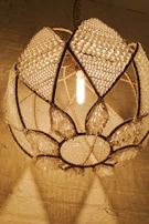 A close-up view of an ornate chandelier with a geometric design, featuring numerous crystal embellishments and beaded textures. The chandelier emits a warm, golden light that casts gentle shadows on a plain plaster wall.