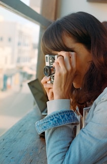 A person is holding a vintage camera to their eye, appearing to take a photo. The setting is near a large window with soft daylight coming through. The person is wearing a light denim jacket and is sitting at a wooden surface with a partially visible laptop nearby.