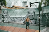 A person is playing basketball on an outdoor court with a tall hoop and a mural featuring two large portraits painted on the wall in the background. The graffiti depicts two men, one wearing a cap and smiling broadly. The urban setting includes high-rise buildings and a fence surrounding the court.