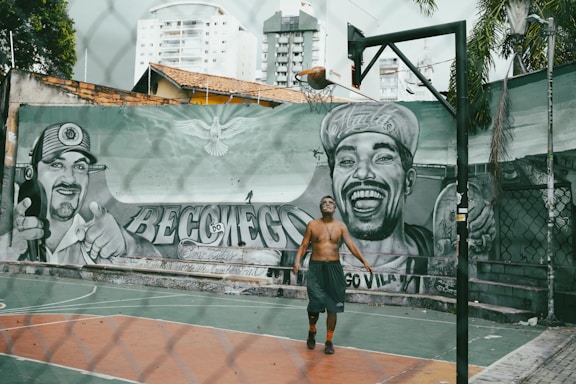 A person is playing basketball on an outdoor court with a tall hoop and a mural featuring two large portraits painted on the wall in the background. The graffiti depicts two men, one wearing a cap and smiling broadly. The urban setting includes high-rise buildings and a fence surrounding the court.