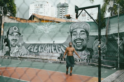 A person is playing basketball on an outdoor court with a tall hoop and a mural featuring two large portraits painted on the wall in the background. The graffiti depicts two men, one wearing a cap and smiling broadly. The urban setting includes high-rise buildings and a fence surrounding the court.