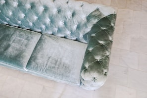 An elegant, plush velvet sofa featuring a quilted design with deep buttoning. The fabric appears to be a soft, muted green with a luxurious texture. The sofa is placed on a smooth, light-colored tiled floor that contrasts with its rich fabric.