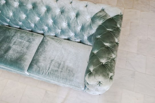 An elegant, plush velvet sofa featuring a quilted design with deep buttoning. The fabric appears to be a soft, muted green with a luxurious texture. The sofa is placed on a smooth, light-colored tiled floor that contrasts with its rich fabric.