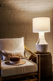 A cozy corner with a woven basket side table supporting a stylish table lamp. The soft light highlights a wooden armchair adorned with a white cushion and a decorative pillow that features intricate patterns. On the chair rests a light brown hat with an ornate band.