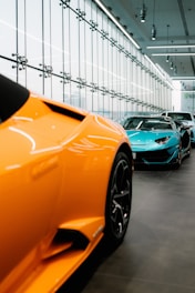 A sleek showroom featuring a lineup of luxury sports cars. The foreground is dominated by an orange car with sharp lines and aerodynamic design, while in the background, a blue car and another vehicle are visible. The space has a spacious modern interior with large glass windows and overhead lighting, creating a streamlined and elegant ambiance.