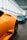 A sleek showroom featuring a lineup of luxury sports cars. The foreground is dominated by an orange car with sharp lines and aerodynamic design, while in the background, a blue car and another vehicle are visible. The space has a spacious modern interior with large glass windows and overhead lighting, creating a streamlined and elegant ambiance.