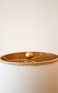 A close-up view of the top of a lampshade with a round, golden finial. The lampshade is a light beige color, and the finial is connected to the lamp's interior framework. The background is a plain, neutral wall.