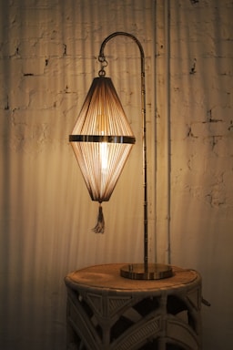 A vintage lamp with a patterned, conical shade exudes warm light, set against a textured white brick wall. It stands on a woven wooden table, creating an ambiance of rustic elegance.