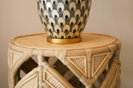 A decorative vase with a patterned design of alternating colors sits atop a round wicker table. The vase has a series of eye-like motifs in black and cream, accented with a golden base. The wicker table shows intricate weaving and natural wood tones.