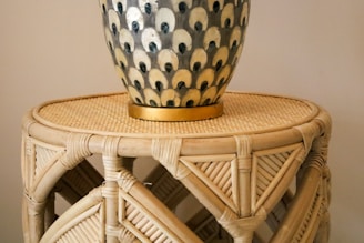 A decorative vase with a patterned design of alternating colors sits atop a round wicker table. The vase has a series of eye-like motifs in black and cream, accented with a golden base. The wicker table shows intricate weaving and natural wood tones.