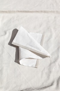 A neatly folded white cloth napkin lies on a textured, light-colored fabric surface. The napkin has a subtle gray border, and soft shadows create a sense of depth.