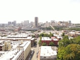 An aerial view of a cityscape with a mix of residential and commercial buildings. High-rise buildings are concentrated in the background, while roads and trees are visible throughout the scene.