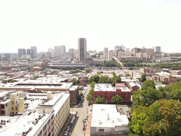 An aerial view of a cityscape with a mix of residential and commercial buildings. High-rise buildings are concentrated in the background, while roads and trees are visible throughout the scene.