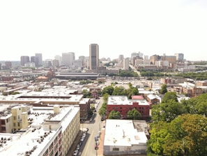 An aerial view of a cityscape with a mix of residential and commercial buildings. High-rise buildings are concentrated in the background, while roads and trees are visible throughout the scene.